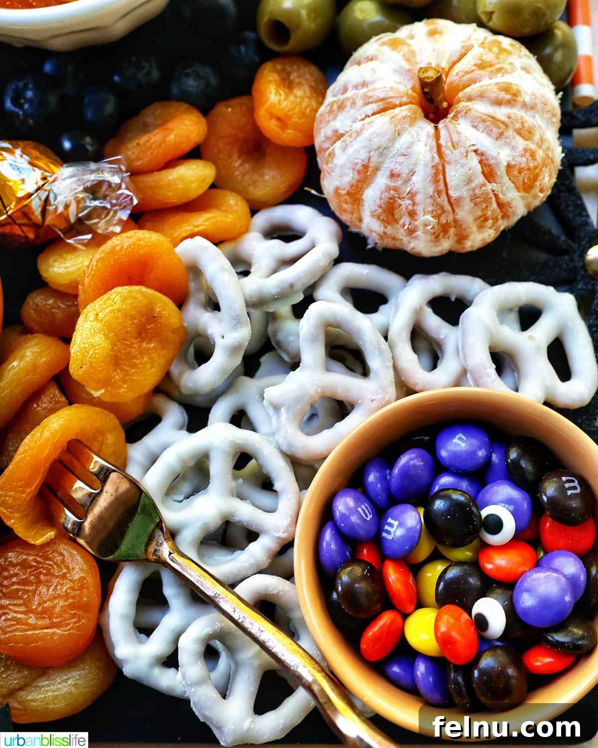 17 Spooktacularly Healthy No-Bake Halloween Treats 5 Close-up image of white chocolate covered pretzels, M&Ms, dried apricots, and oranges arranged on a Halloween charcuterie board.