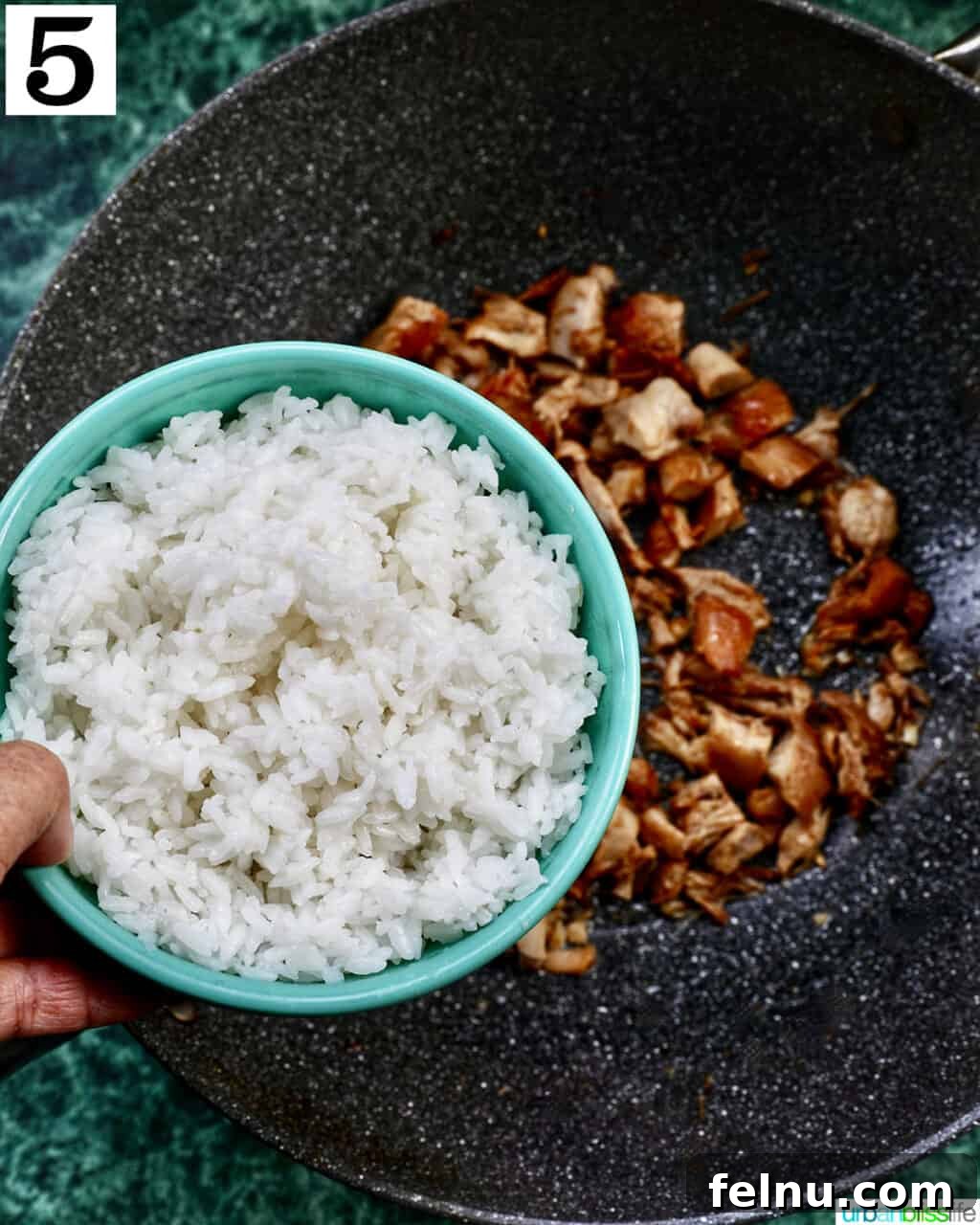 Savory Garlic Adobo Fried Rice 9 A hand adding rice from a bowl into a wok containing adobo chicken, ready for stir-frying.