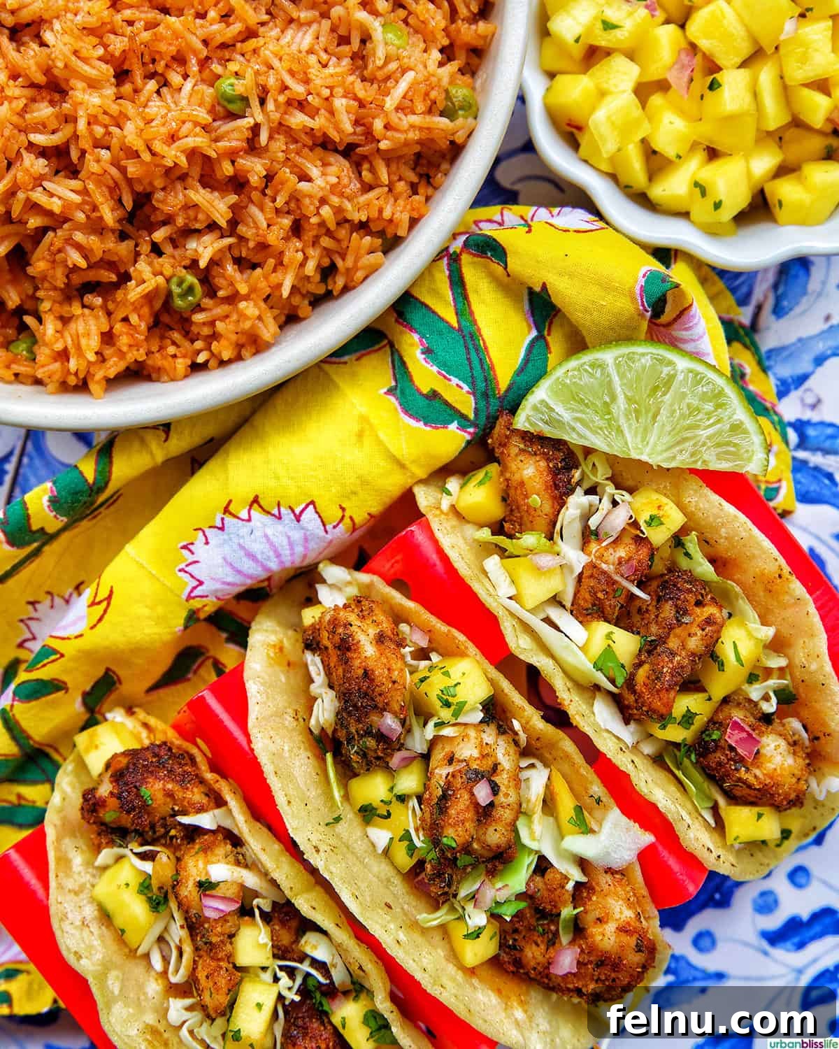 Three air fryer shrimp tacos in a red taco stand, accompanied by a side of Mexican rice and fresh mango salsa for a complete meal.